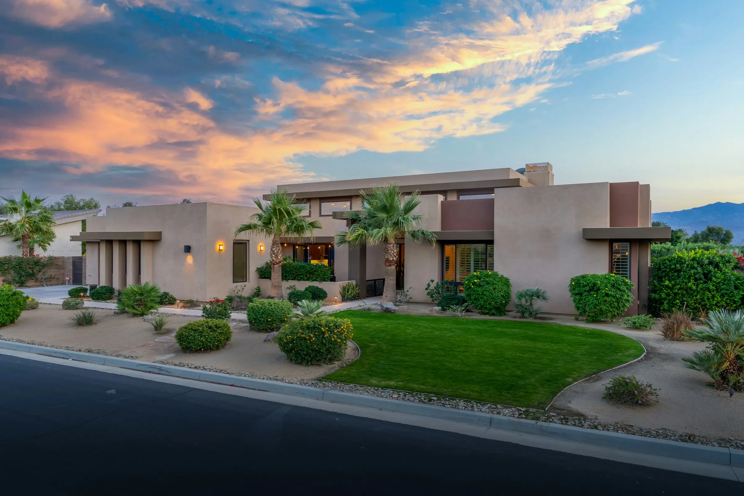 Single-story desert home at dusk with trimmed shrubs, gravel beds, palms, and a small lawn
